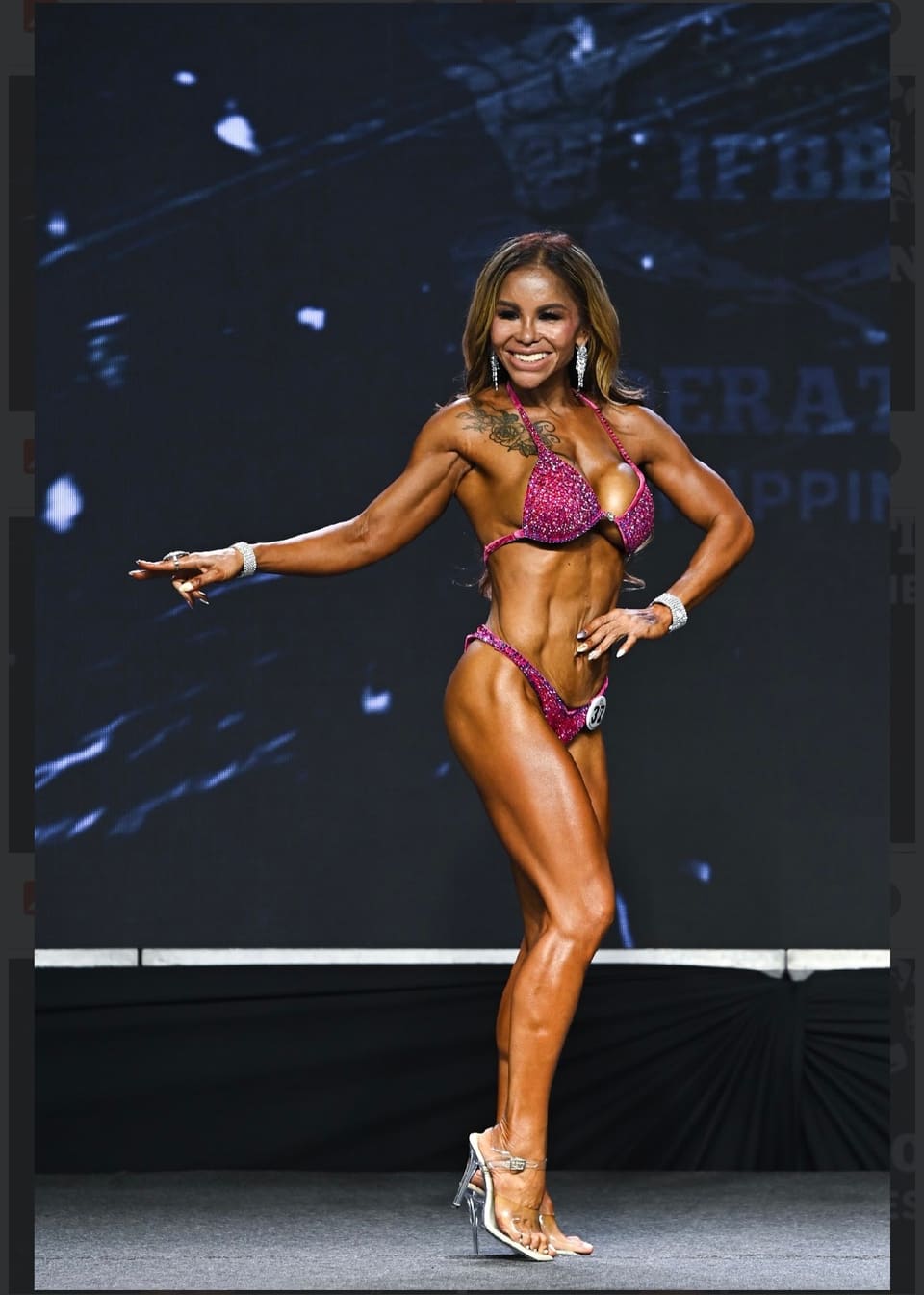 Coach Jasmin Rough posing on a dark competition stage in a pink bikini